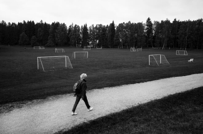 A person walks on a gravel path beside a field with 10 football goals. Behind the field is a stand of trees.